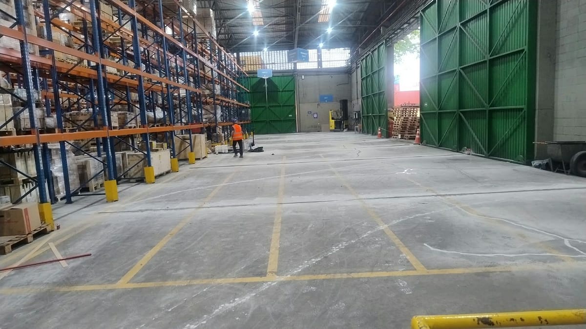 Armazém com estruturas porta-paletes carregadas e piso de concreto em recuperação, com a frente de trabalho ao fundo