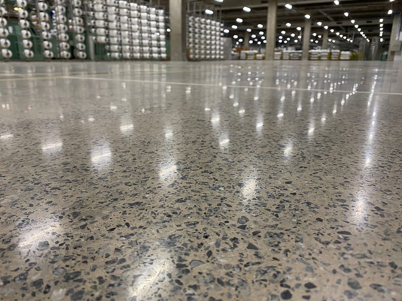 Detalhe rente ao chão de piso de concreto lapidado com agregado exposto em indústria têxtil, luminárias refletidas até o fundo do galpão