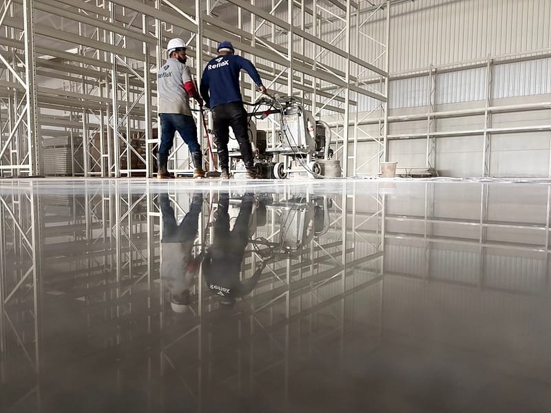 Dois operadores da Reflex conduzem a politriz planetária em galpão com porta-paletes, com o piso de concreto lapidado refletindo a estrutura