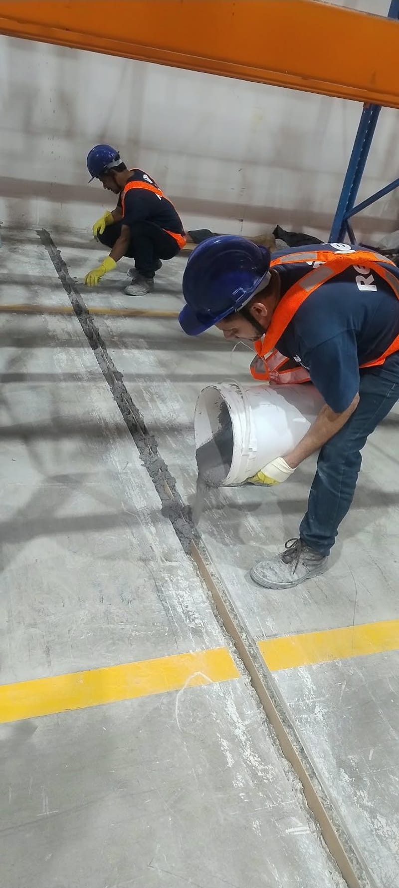 Dois profissionais de capacete e colete trabalham em uma junta aberta do piso, um deles vertendo o material, em armazém com porta-paletes