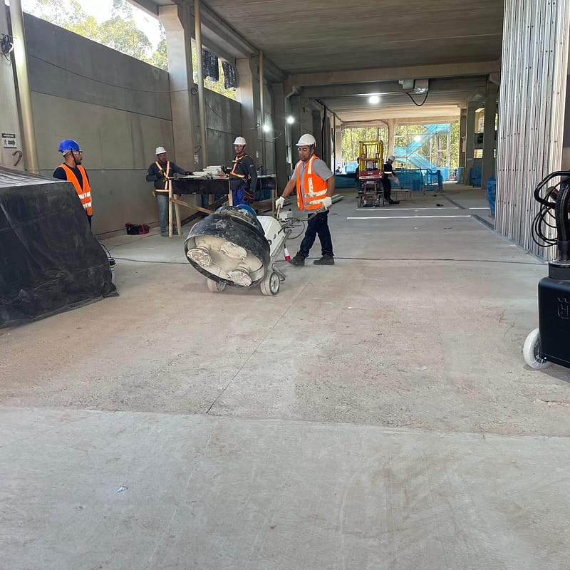 Equipe com capacetes e coletes em torno da politriz, na preparação mecânica do piso de concreto em edifício em obra