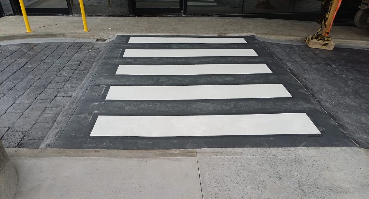 Faixa de pedestre recém pintada, com listras brancas sobre fundo cinza escuro, no piso de concreto estampado de uma área coberta de acesso