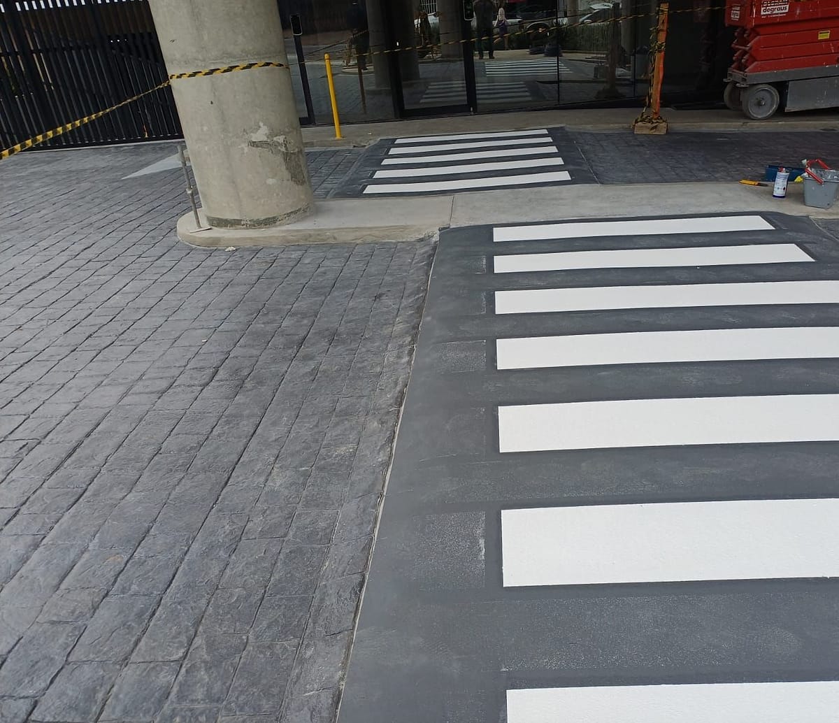 Duas faixas de pedestre pintadas no piso de concreto estampado, entre pilares, diante da entrada envidraçada de um prédio comercial