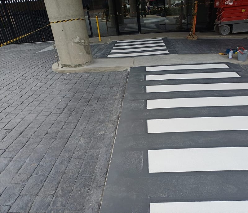 Duas faixas de pedestre pintadas no piso de concreto estampado, entre pilares, diante da entrada envidraçada de um prédio comercial