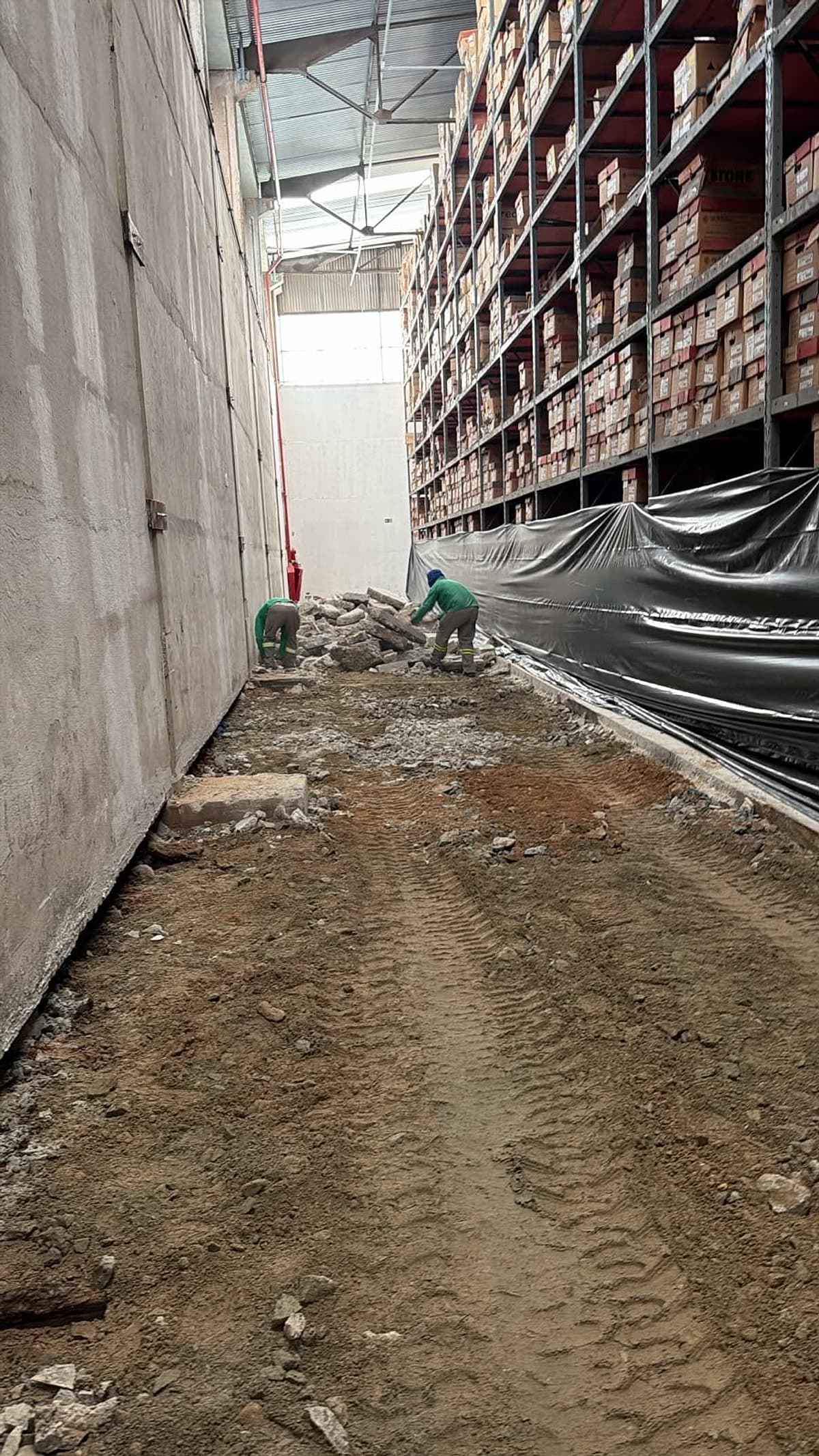 Faixa de piso demolida junto à parede externa do armazém, com a sub-base exposta e as estruturas de armazenagem cobertas por lona preta