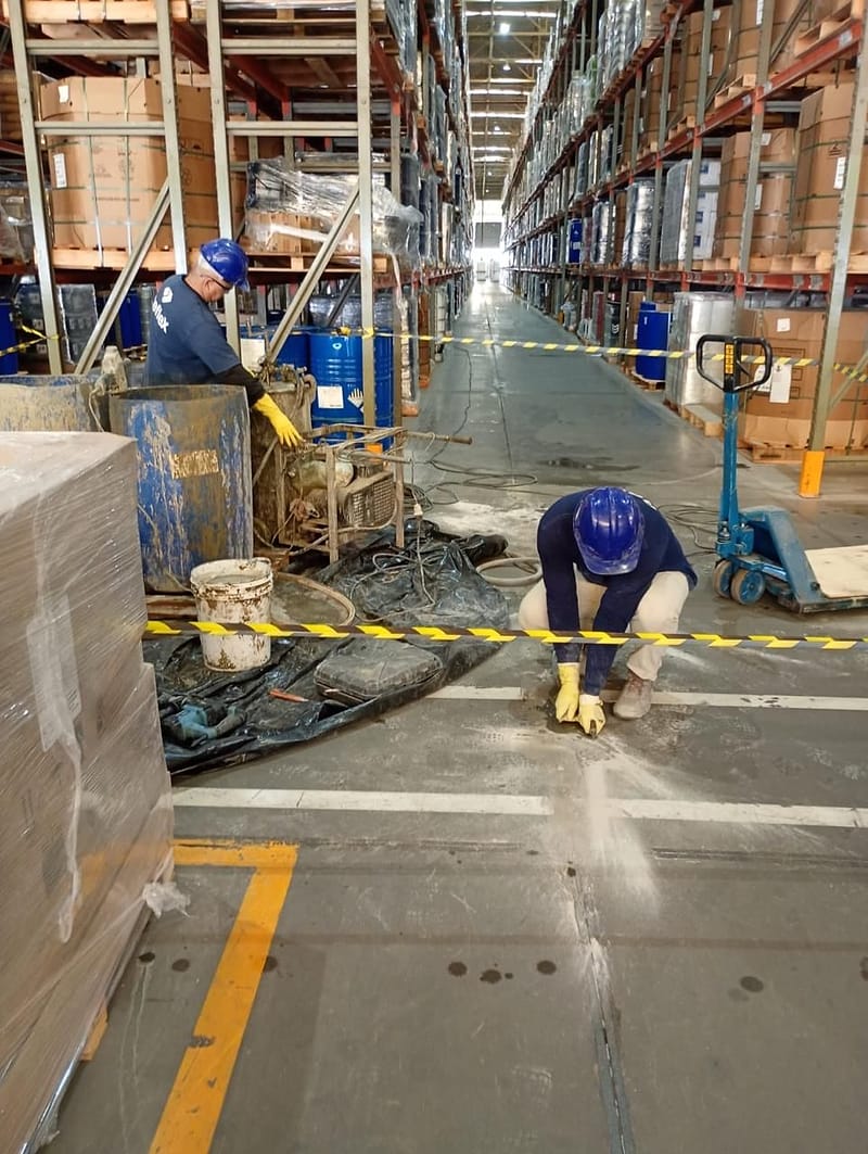 Técnico da Reflex injeta calda de cimento em furo junto à junta do piso de concreto, no corredor de um armazém com porta-paletes em operação