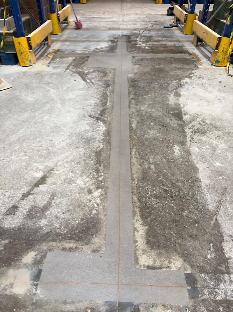 Cruzamento de juntas recuperadas no corredor entre porta-paletes, com as faixas de reparo novas cortando o piso de concreto desgastado