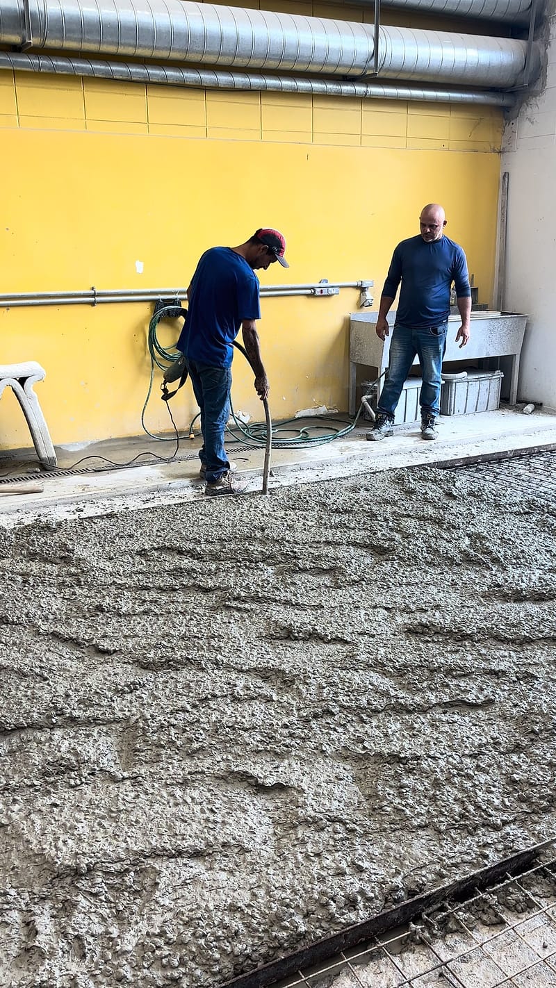 Dois operadores lançam e espalham concreto fresco em um trecho de piso industrial junto à parede