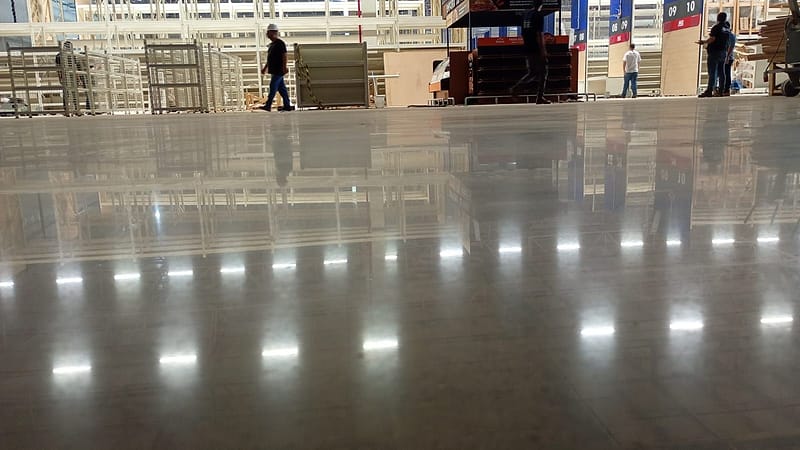 Piso de concreto lapidado de brilho uniforme em área de vendas de atacarejo, com equipes de montagem circulando ao fundo