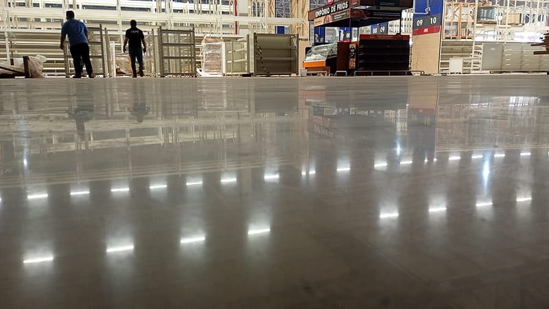 Vista rente ao chão de piso de concreto lapidado em loja de atacarejo, refletindo gôndolas vazias e fileiras de luminárias