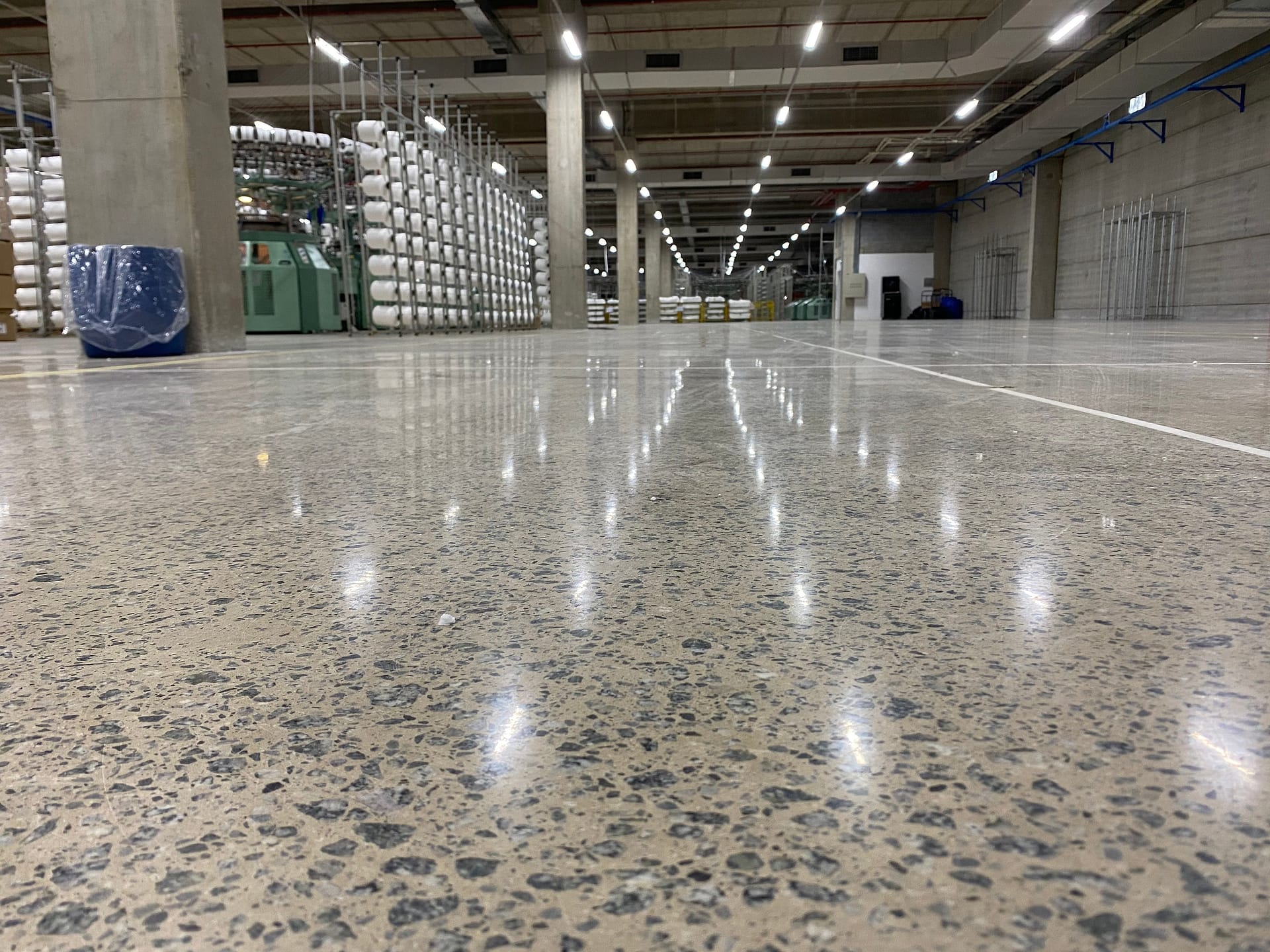 Piso de concreto lapidado de alto brilho em fábrica têxtil de grande área, refletindo dezenas de luminárias entre pilares e máquinas