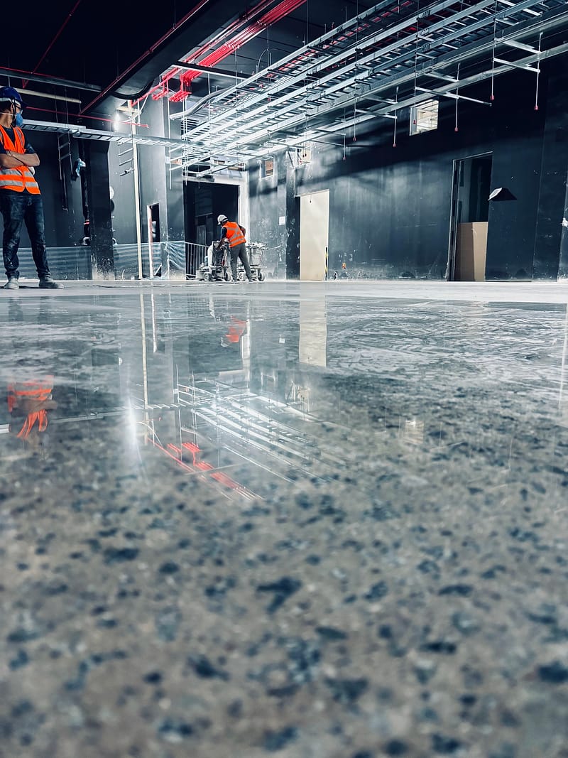 Operador de colete refletivo conduz a politriz sobre piso de concreto lapidado escuro, com o reflexo do galpão e dos equipamentos na superfície