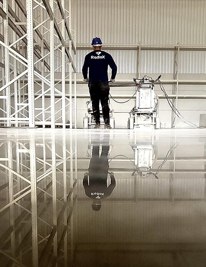 Técnico da Reflex de frente para a politriz em galpão, com o corpo inteiro refletido no piso de concreto espelhado