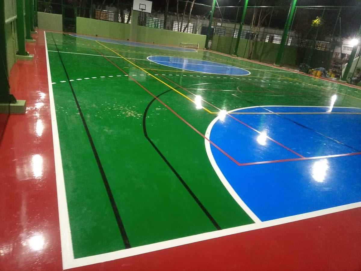Quadra poliesportiva coberta com piso pintado em verde, azul e vermelho e demarcação de futsal, vôlei e basquete