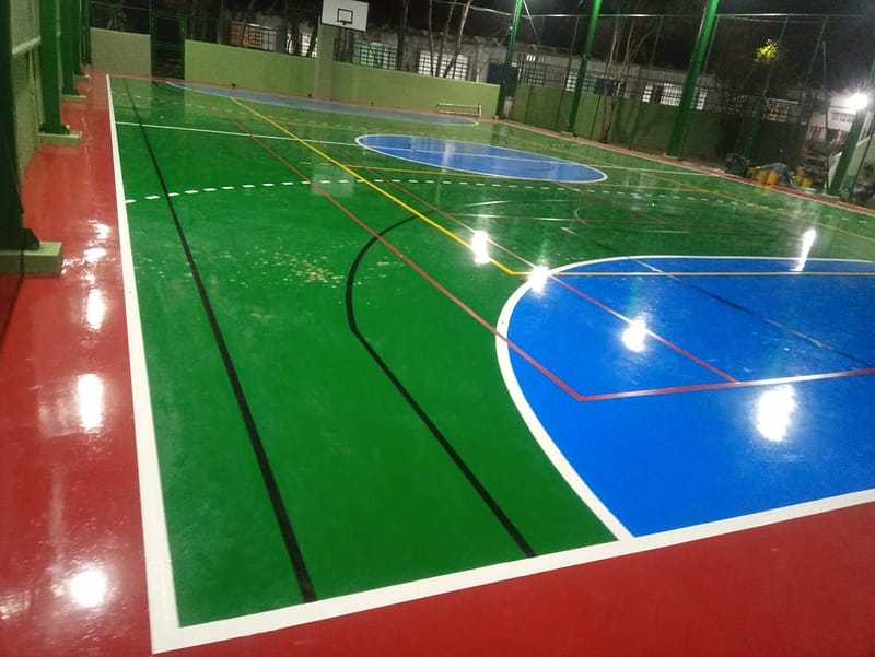 Quadra poliesportiva coberta com piso pintado em verde, azul e vermelho e demarcação de futsal, vôlei e basquete