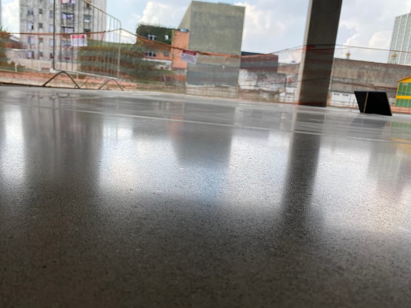 Piso de concreto lapidado em galpão aberto, refletindo a luz do dia e as fachadas do entorno