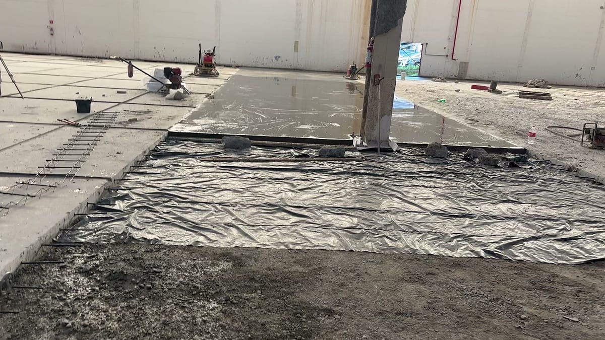 Trecho de piso de concreto recém concretado junto a um pilar, coberto com lona plástica para a cura, com a base preparada ao lado