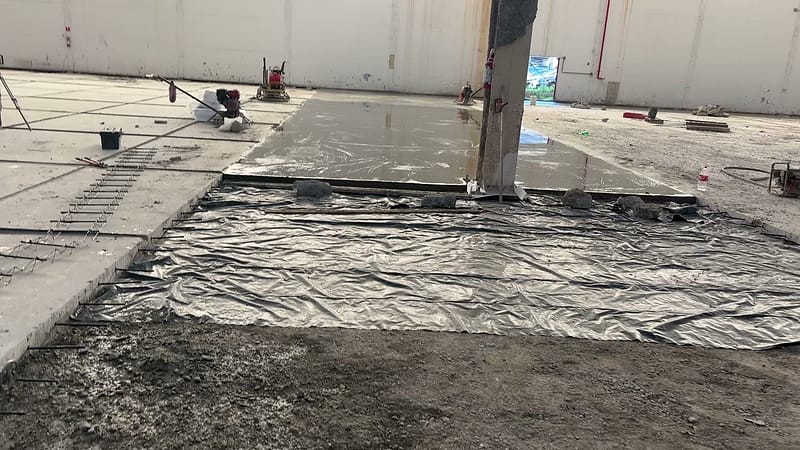 Trecho de piso de concreto recém concretado junto a um pilar, coberto com lona plástica para a cura, com a base preparada ao lado