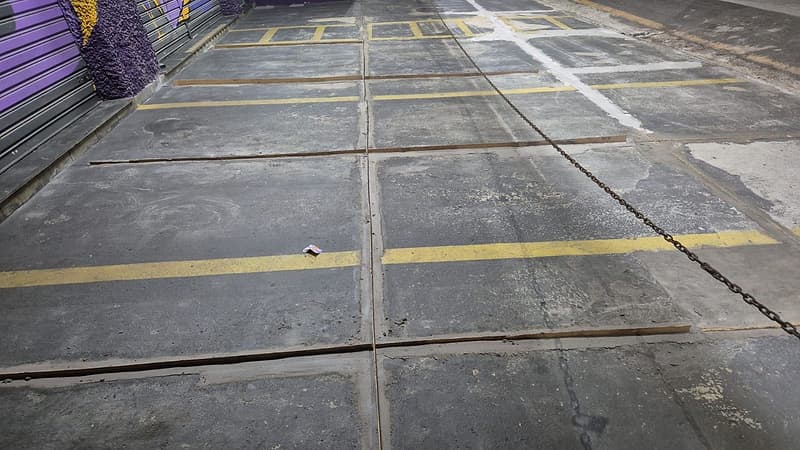 Estacionamento frontal de loja com placas de concreto escurecidas e juntas abertas, isolado por corrente à noite