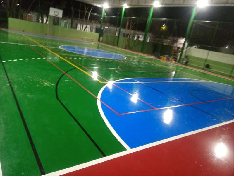 Garrafão de basquete pintado de azul sobre quadra verde brilhante, sob refletores, em quadra coberta