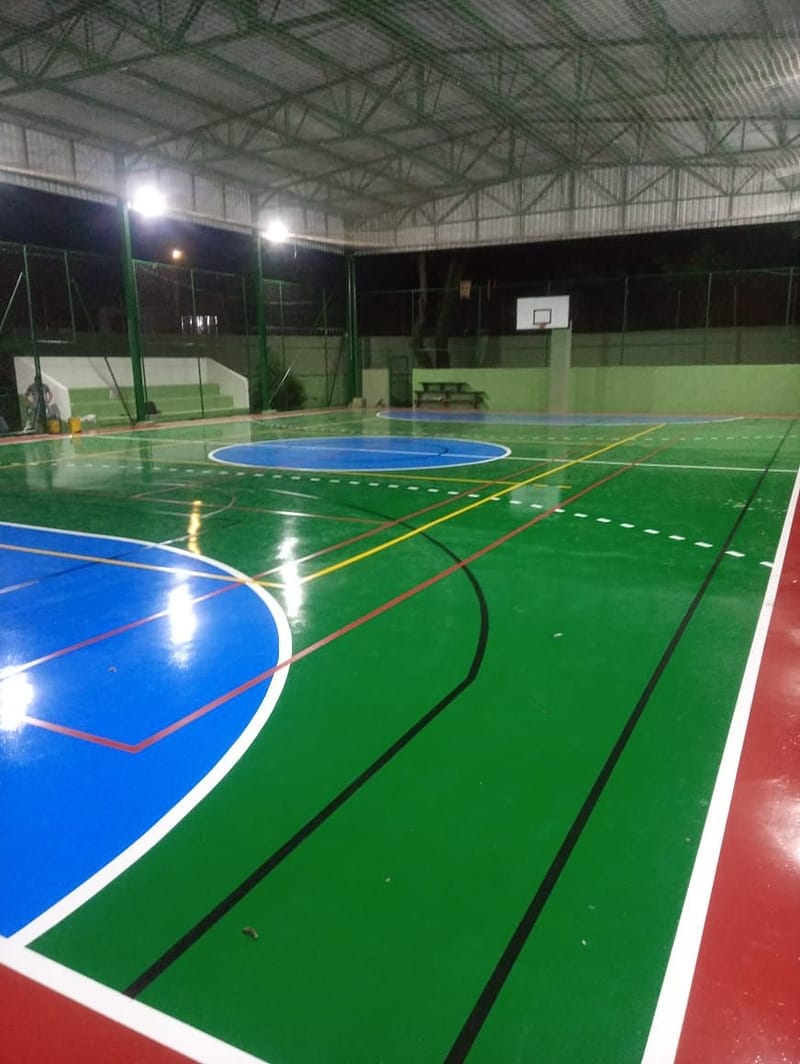 Vista geral de quadra poliesportiva coberta com demarcação de futsal, vôlei e basquete e tabela ao fundo