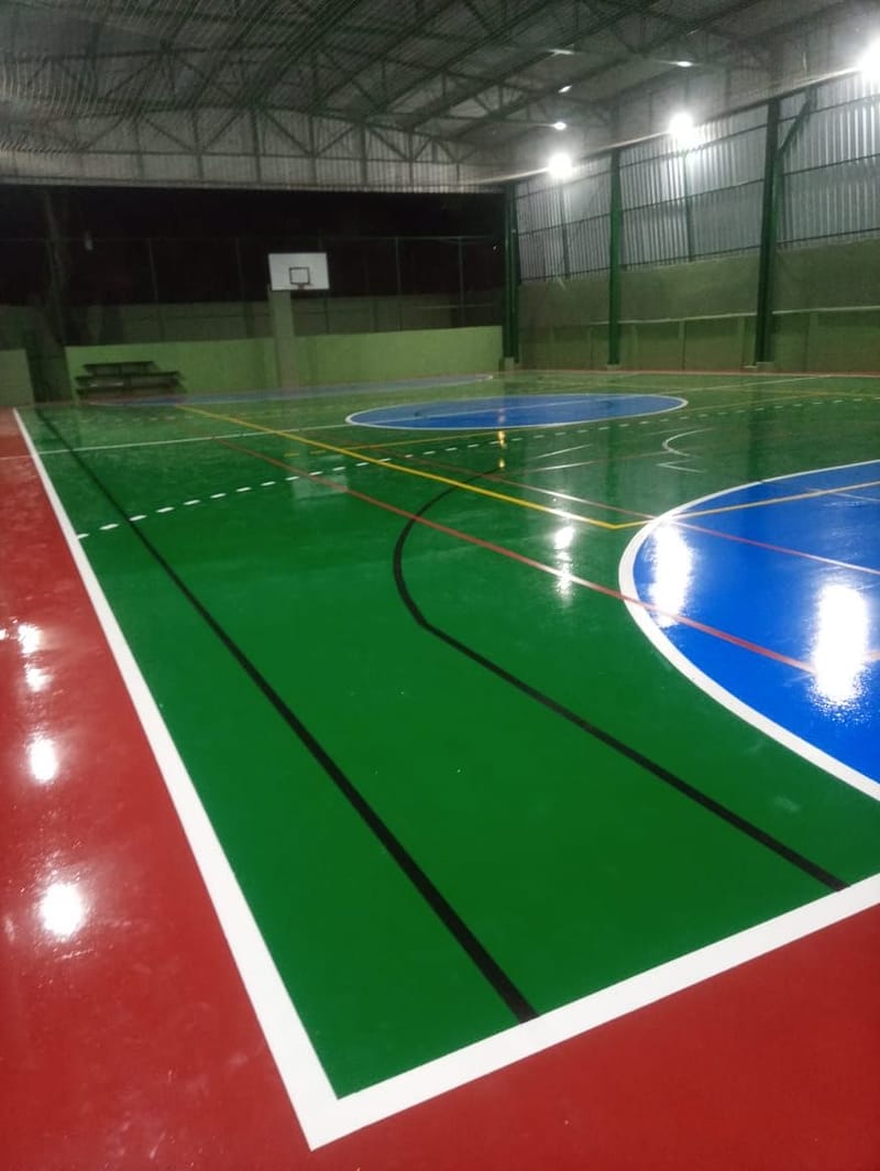 Canto de quadra poliesportiva com linha branca larga separando o campo verde do recuo vermelho