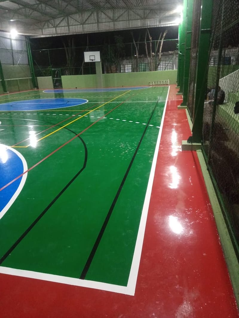 Lateral de quadra poliesportiva com recuo vermelho junto ao alambrado e tabela de basquete ao fundo